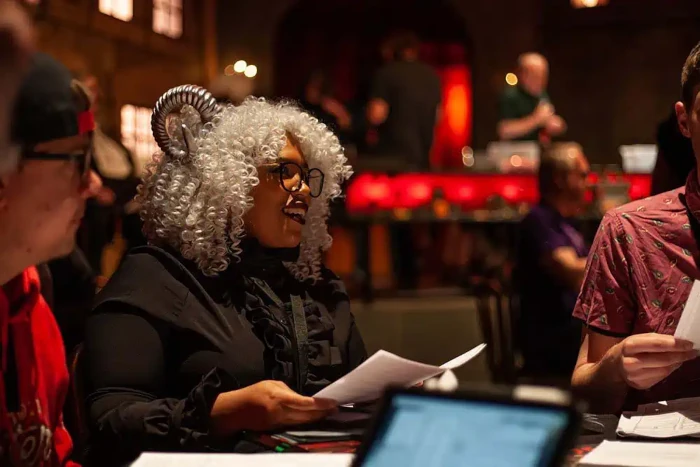 A close-up on a gaming table focuses on a Black woman in a black frilly blouse with white curly should-length hair, black-rimmed glasses, and silver horns on her head smiling at someone off-screen while holding sheets of paper. 